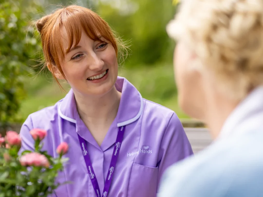 A smiling woman in a purple uniform with a Helping Hands logo talks to another person outdoors, near pink flowers and greenery.