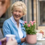 An older woman with short blonde hair, wearing a blue cardigan, smiles while holding a biscuit and sitting at an outdoor table with tea and pink flowers, chatting with another person.