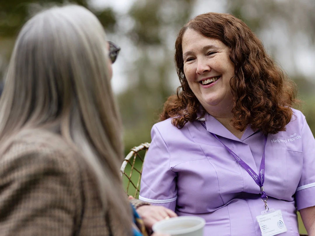 A woman in a lavender uniform with a name badge and lanyard smiles warmly at an older woman with long grey hair during an outdoor conversation.