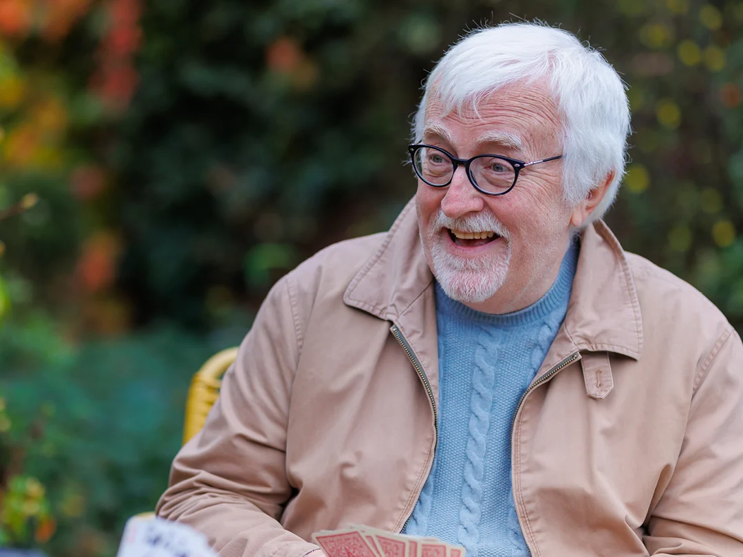 An older man with white hair and glasses, wearing a beige jacket and blue jumper, smiles whilst holding playing cards outdoors. The background is blurred with green foliage.