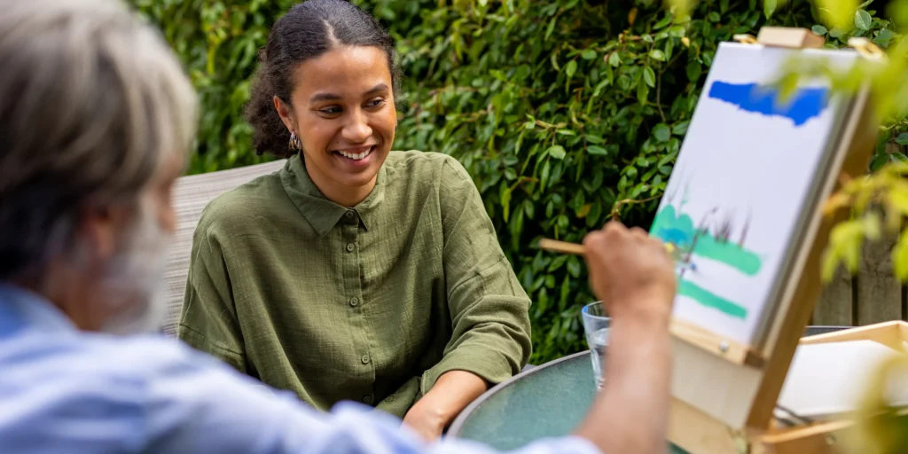 A woman smiles whilst sitting at a table outdoors, watching another person paint on a canvas placed on an easel. Green plants form the background, offering a peaceful setting to discuss how carers can support New Year’s resolutions.