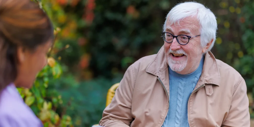 An elderly man with white hair, glasses, and a beard smiles whilst talking to a woman outdoors, with greenery and colourful foliage in the background.