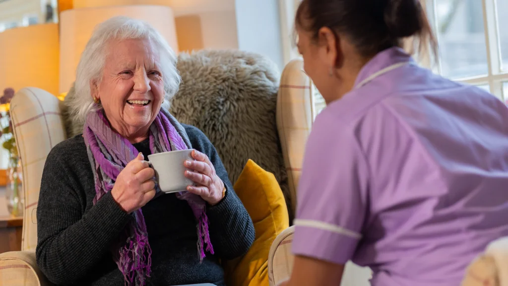 An elderly woman sits in a cosy armchair, smiling and holding a mug, whilst chatting with a carer in a purple uniform. Warm lighting and soft furnishings create a comfortable, welcoming atmosphere.
