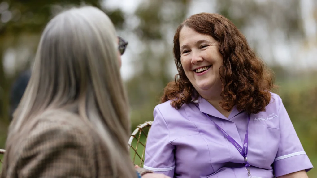 A woman in a lavender uniform smiles warmly whilst talking to another person with long grey hair outdoors. The atmosphere is friendly and relaxed.