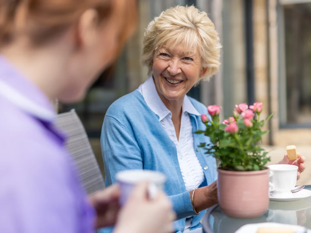An older woman in a blue cardigan smiles whilst having tea and biscuits at an outdoor table with another person. A pink potted flower and two cups are on the table.