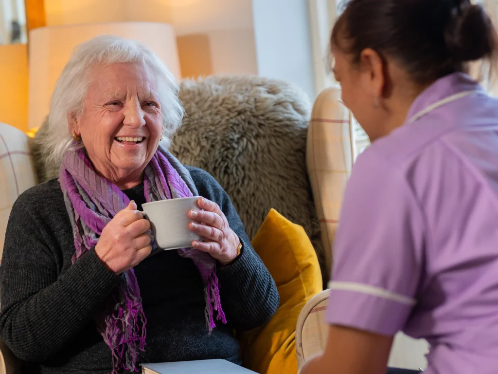An elderly woman with grey hair, wearing a purple scarf, smiles and holds a mug while sitting on an armchair, talking to a carer in a cosy, well-lit room.