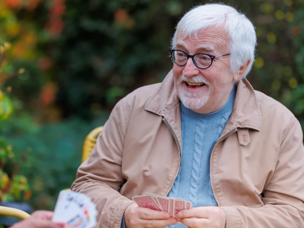 An older man with white hair and glasses smiles whilst playing cards outdoors. He is wearing a light beige jacket and a blue jumper. Another person’s hand holding cards is visible in the foreground.