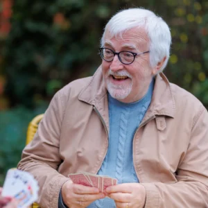 An older man with white hair and glasses, smiling and holding playing cards, sits outdoors wearing a beige jacket and blue jumper. Another person holding cards is partially visible in the foreground.