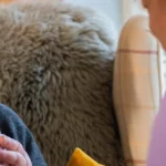 An elderly woman smiles whilst holding a mug, sitting in a cosy chair with a purple scarf around her neck, as she talks with a carer in a light purple uniform.