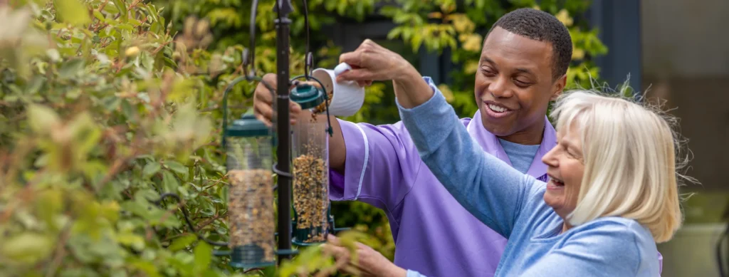 A smiling man and woman fill bird feeders with seeds in a garden, surrounded by green foliage. The man pours seeds whilst the woman holds another feeder, both appearing cheerful and engaged in the activity.