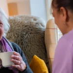 An elderly woman sits in an armchair, smiling and holding a mug, whilst talking to a carer in a purple uniform. The setting appears warm and cosy, with soft lighting and homely decor.