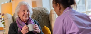 An elderly woman sits in an armchair, smiling and holding a mug, whilst talking to a carer in a purple uniform. The setting appears warm and cosy, with soft lighting and homely decor.