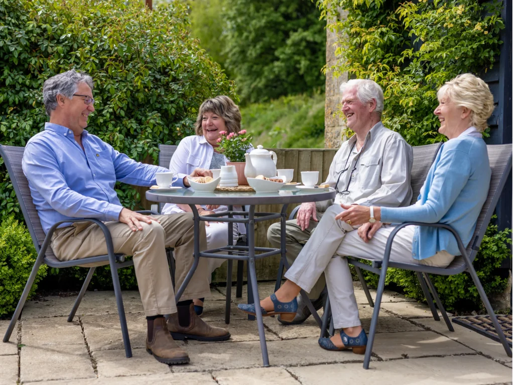 Four older adults sit around an outdoor table, smiling and enjoying tea and snacks on a sunny patio, surrounded by lush green plants and trees.