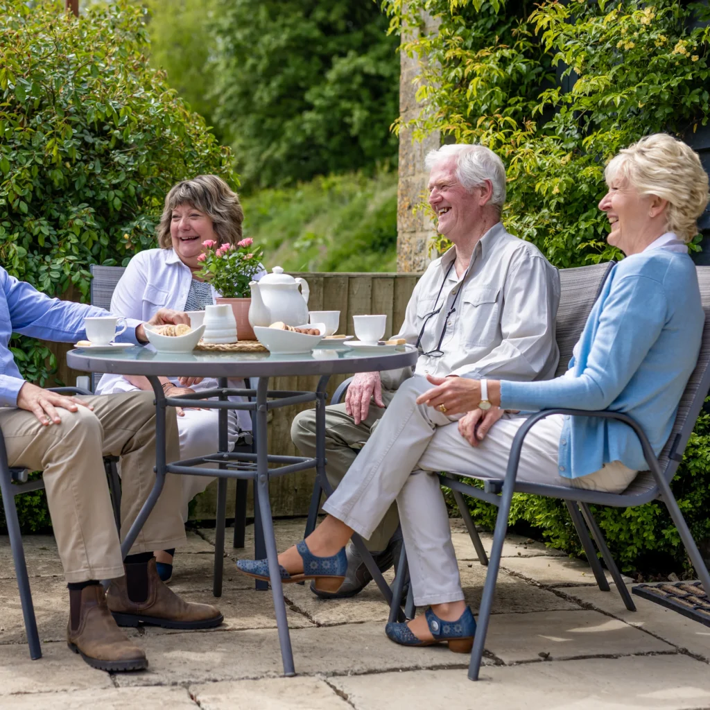 Four older adults sit around a patio table outdoors, smiling and laughing as they enjoy tea and biscuits together. The setting is bright and lush with greenery in the background.