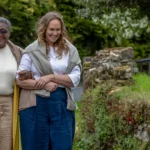Three women walk outdoors on a path, smiling and arm-in-arm, surrounded by greenery and trees. They appear relaxed and are dressed in casual, comfortable clothing.
