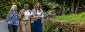 Three women walk outdoors on a path, smiling and arm-in-arm, surrounded by greenery and trees. They appear relaxed and are dressed in casual, comfortable clothing.