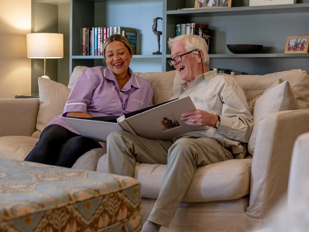 A smiling elderly man and a female carer sit together on a sofa, looking at a large photo album. Bookcases, books, and framed photos are visible in the background, creating a cosy and warm atmosphere.
