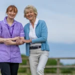 A smiling carer in a purple uniform stands outdoors, linking arms with an older woman in a light blue cardigan and white blouse. They both look happy, with greenery and a wooden fence in the background.