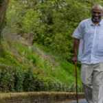 An elderly man with a walking stick walks outdoors on a leafy path, smiling, while being assisted by a younger man in a purple uniform. They appear happy and engaged in conversation.