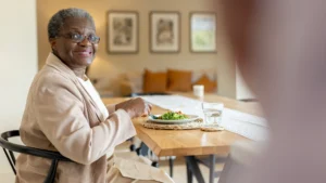 An older woman wearing glasses and a beige jacket sits at a wooden table, smiling at the camera. She is eating a meal with vegetables and has a glass of water in front of her in a cosy, well-lit room.