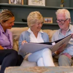 Three people sit on a sofa smiling and looking at a photo album together. An elderly man holds the album while an elderly woman and a younger woman in a purple uniform enjoy the moment. Shelves with framed photos are in the background.