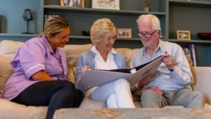 Three people sit on a sofa smiling and looking at a photo album together. An elderly man holds the album while an elderly woman and a younger woman in a purple uniform enjoy the moment. Shelves with framed photos are in the background.