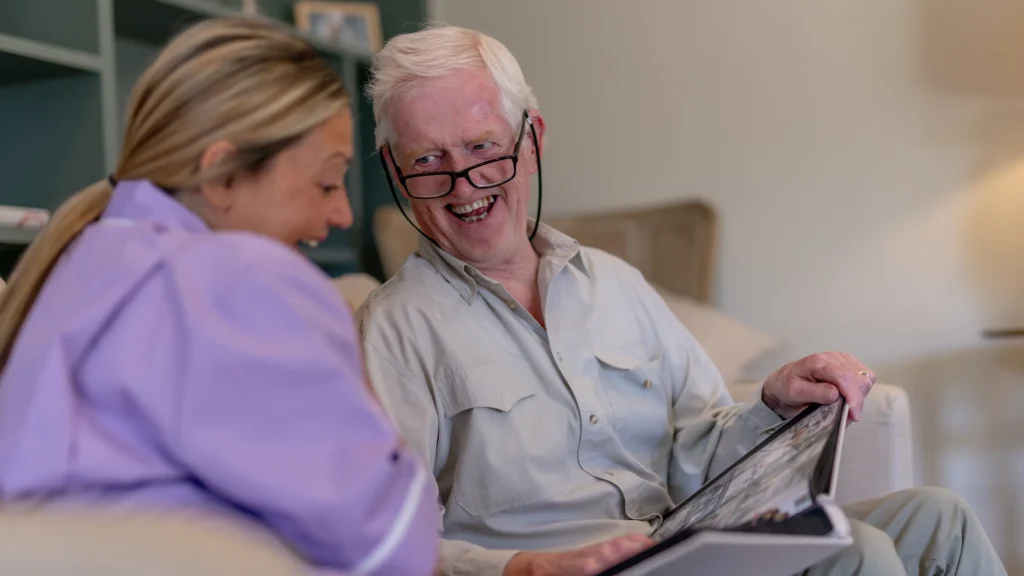 An elderly man with glasses smiles and holds a photo album while sitting on a sofa with a woman. Both are looking at the album and appear to be enjoying a pleasant moment together.