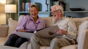 An elderly man and a carer sit on a sofa, smiling and looking at a large photo album together. Bookshelves and a lamp are visible in the background, suggesting a cosy living room setting.