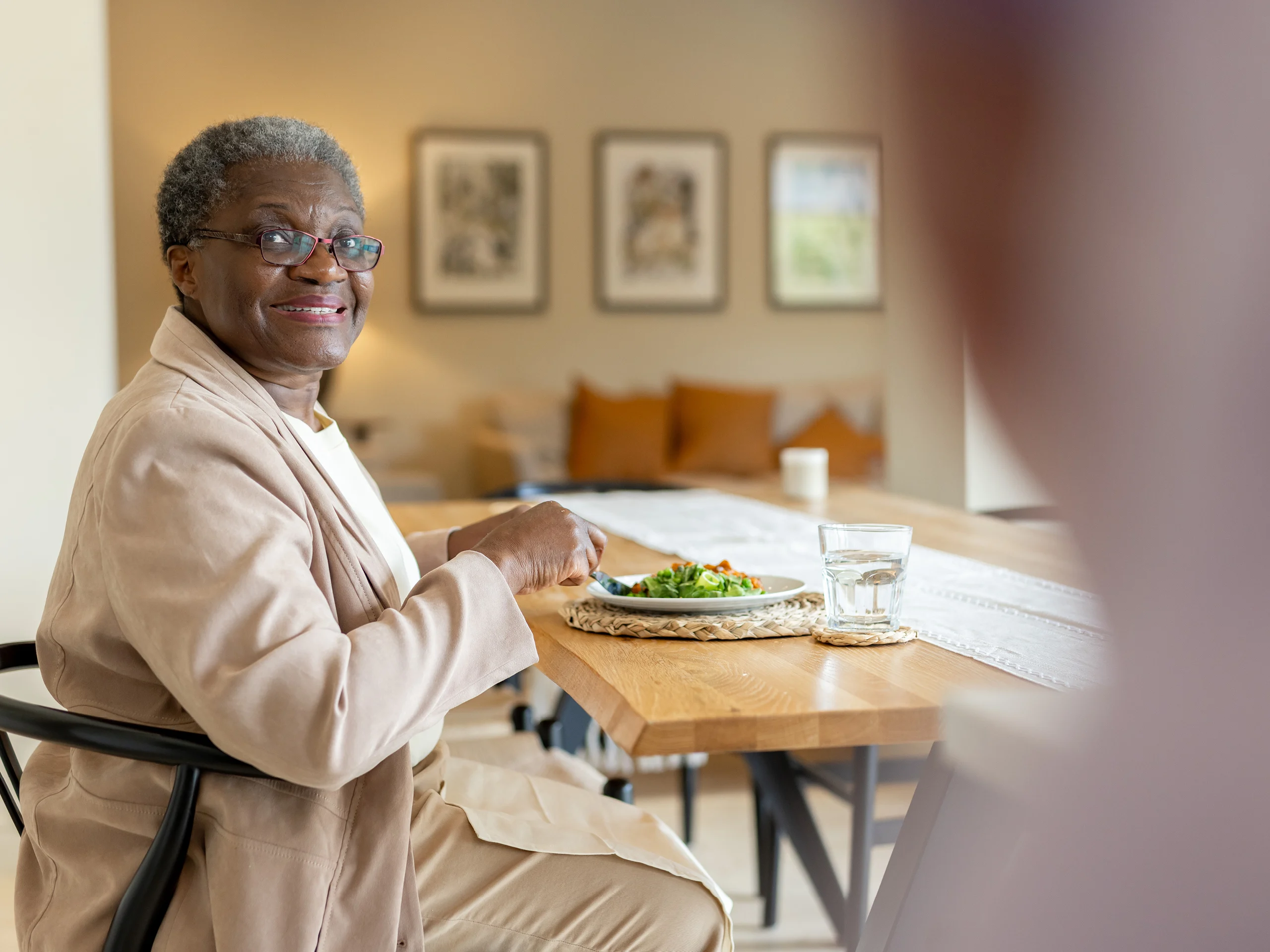 An older woman wearing glasses and a beige jacket sits at a wooden table, smiling at the camera. She is eating a meal with a glass of water, in a cosy, well-lit room with framed art on the wall.