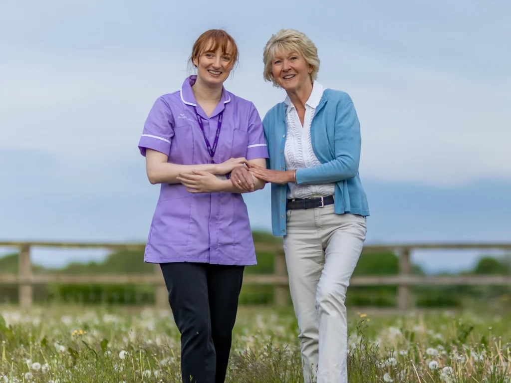A smiling elderly woman walks arm-in-arm with a carer in a purple uniform across a grassy field with wildflowers, under a clear sky.