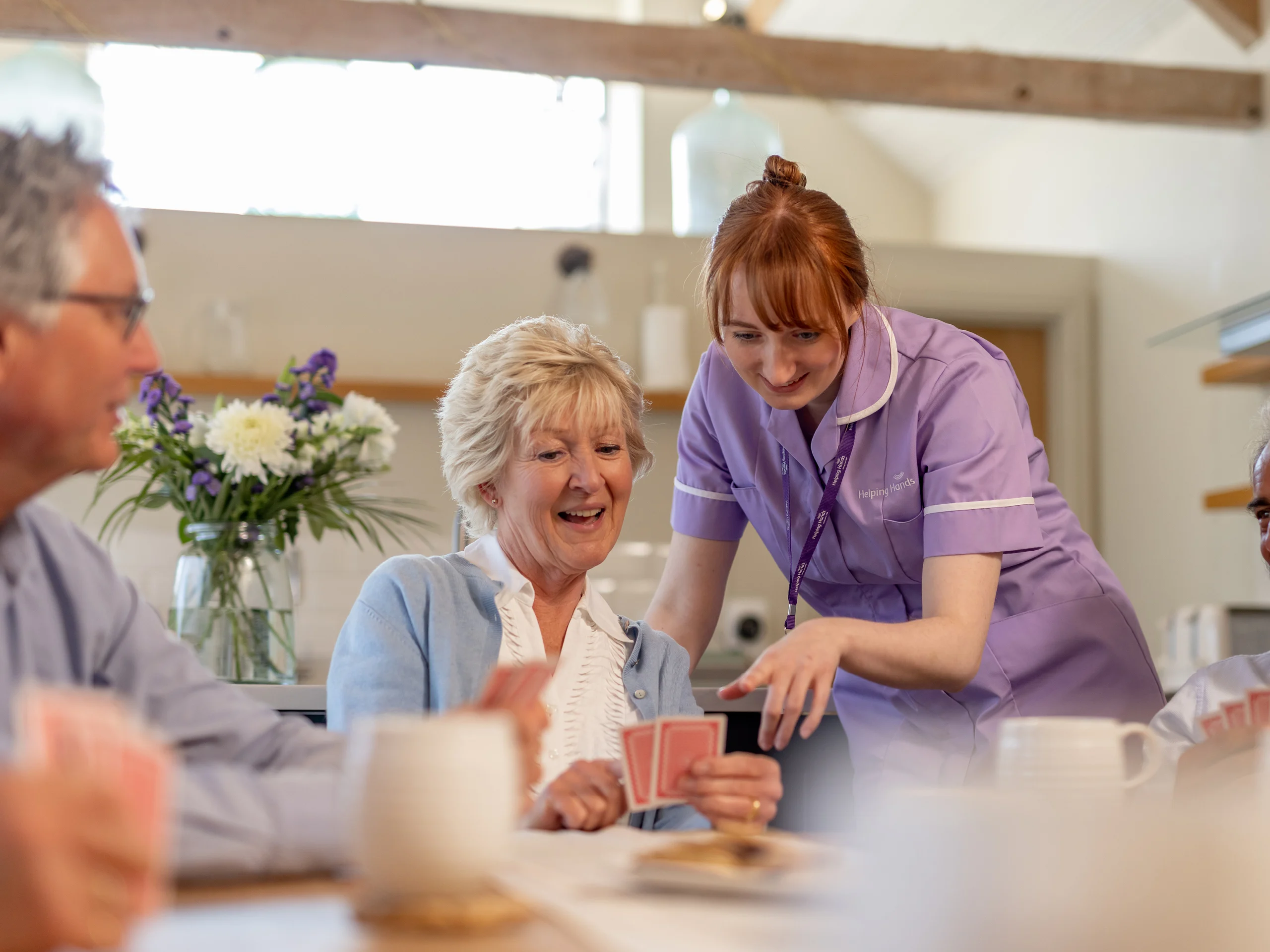 A smiling elderly woman holds playing cards while a carer in a purple uniform leans over and points, helping her during a card game at a bright, cosy table with others.