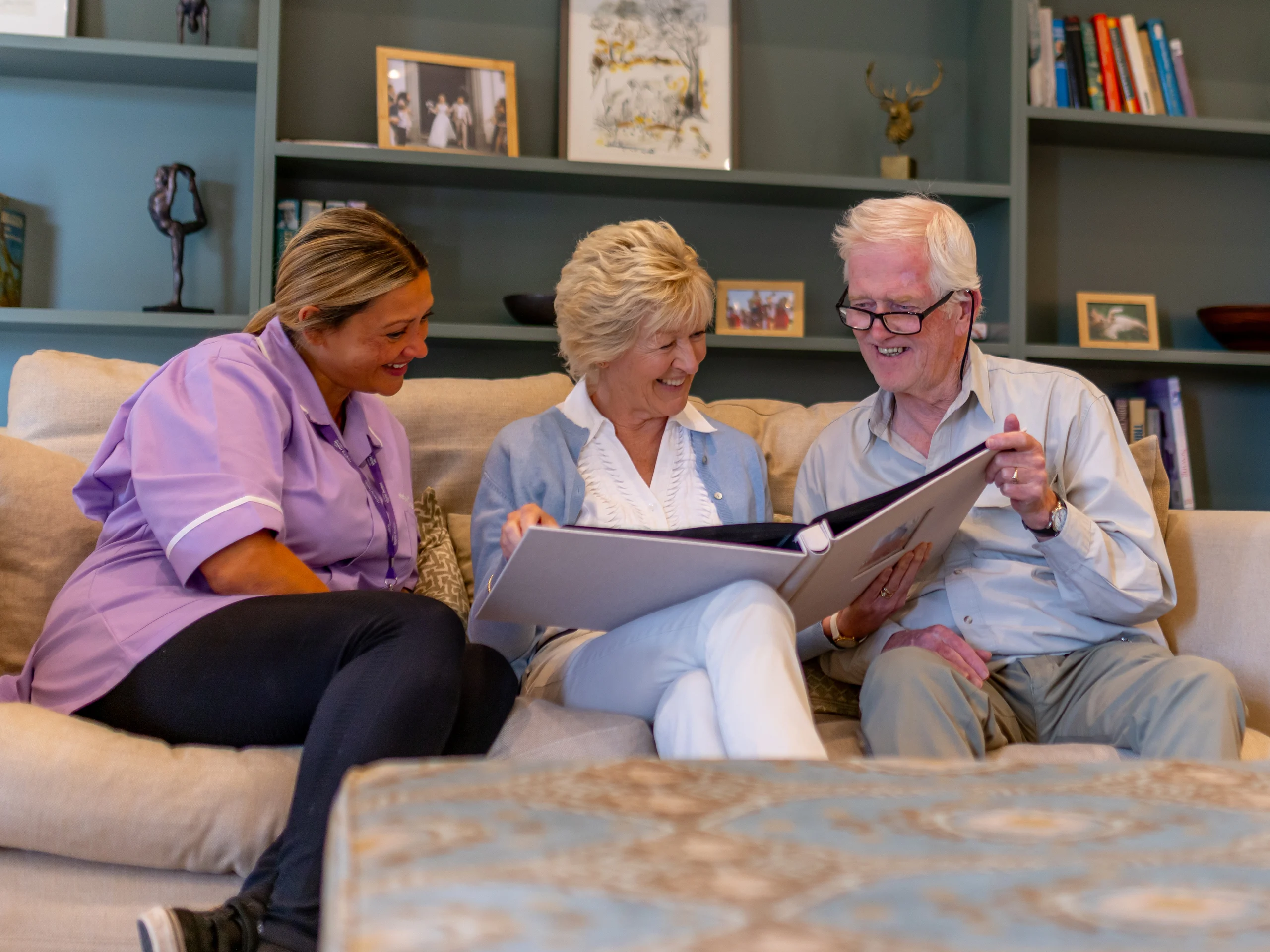Three people sit on a sofa, smiling and looking at a large photo album together. Two are older adults, and one appears to be a carer. The background shows shelves with books and framed photographs.