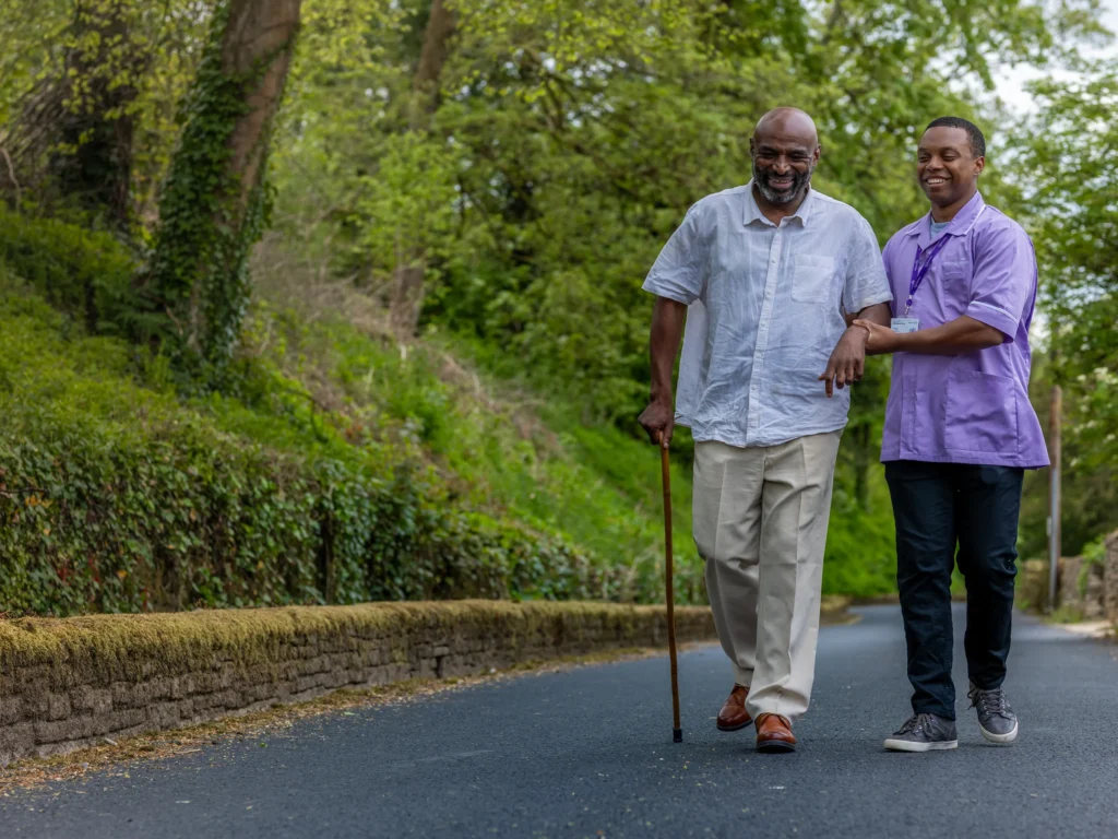 An elderly man with a walking stick walks outdoors on a path, smiling and holding the arm of a younger man in a purple shirt who is assisting him. Trees and greenery line the background.