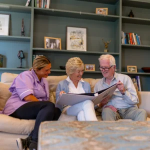 Three elderly people sit on a sofa, smiling and looking at a large photo album together, in a cosy living room with shelves and framed pictures in the background.