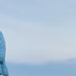 A smiling carer in a purple uniform stands outdoors next to an older woman in a light blue cardigan, holding her arm, with a blue sky in the background.