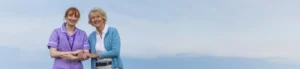 A smiling carer in a purple uniform stands outdoors next to an older woman in a light blue cardigan, holding her arm, with a blue sky in the background.