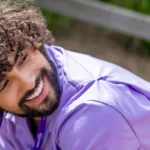 A young man with curly hair smiles warmly at an older man as they sit and talk outdoors, surrounded by greenery.