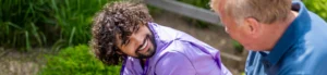 A young man with curly hair smiles warmly at an older man as they sit and talk outdoors, surrounded by greenery.