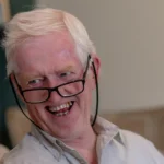 An older man with white hair and glasses smiles warmly at a woman with blonde hair, both engaged in a pleasant conversation in a cosy indoor setting.