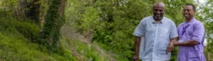 Two men walking outdoors in a lush, green setting. One man, wearing a white shirt, uses a walking stick and is being supported by the other man, who is wearing a light purple uniform and smiling.