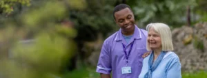A smiling healthcare worker in a purple uniform stands outdoors with an elderly woman, gently placing a hand on her shoulder as they both smile, surrounded by greenery.