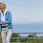 A smiling carer in a purple uniform supports an older woman in a blue cardigan as they stand together outdoors, with a wooden fence and greenery in the background.