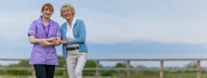 A smiling carer in a purple uniform supports an older woman in a blue cardigan as they stand together outdoors, with a wooden fence and greenery in the background.