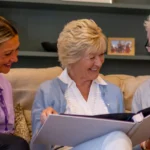 Three adults sit on a sofa smiling and looking at a large photo album together; an elderly couple sits with a carer in a purple uniform, in a cosy living room with bookshelves in the background.