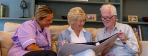 Three adults sit on a sofa smiling and looking at a large photo album together; an elderly couple sits with a carer in a purple uniform, in a cosy living room with bookshelves in the background.