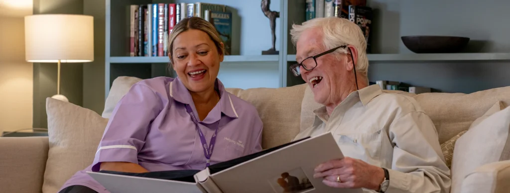 A smiling elderly man and a carer in a purple uniform sit on a sofa, looking at a photo album together. Bookcases and a lamp are visible in the background.