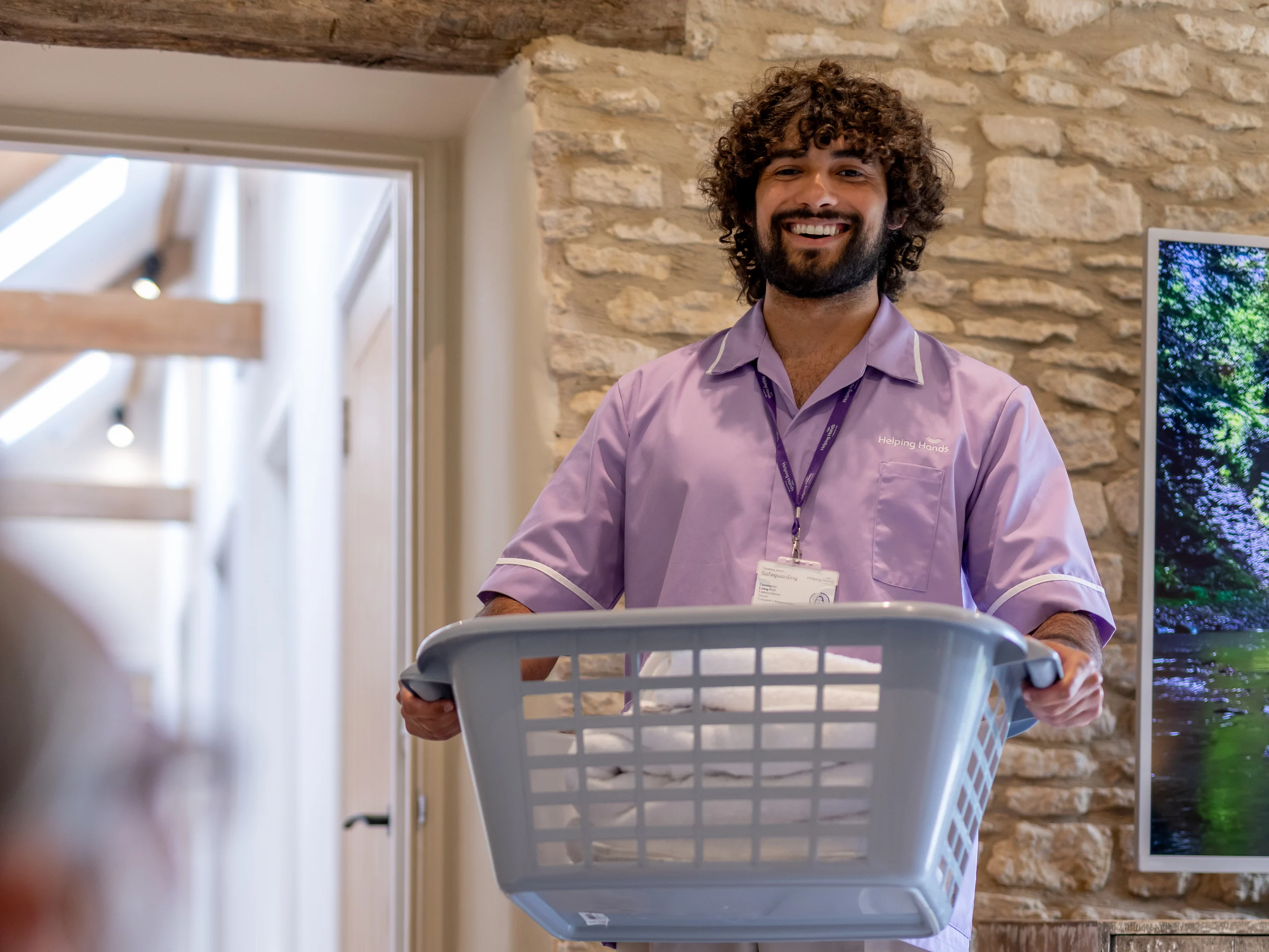 A smiling man with curly hair, wearing a light purple uniform and a lanyard, holds a laundry basket filled with white washing in a cosy, stone-walled room.