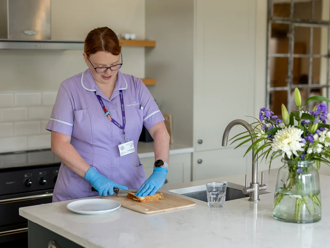 A woman in a light purple uniform and blue gloves slices bread on a chopping board in a modern kitchen. A vase of flowers and a glass of water sit on the worktop nearby.