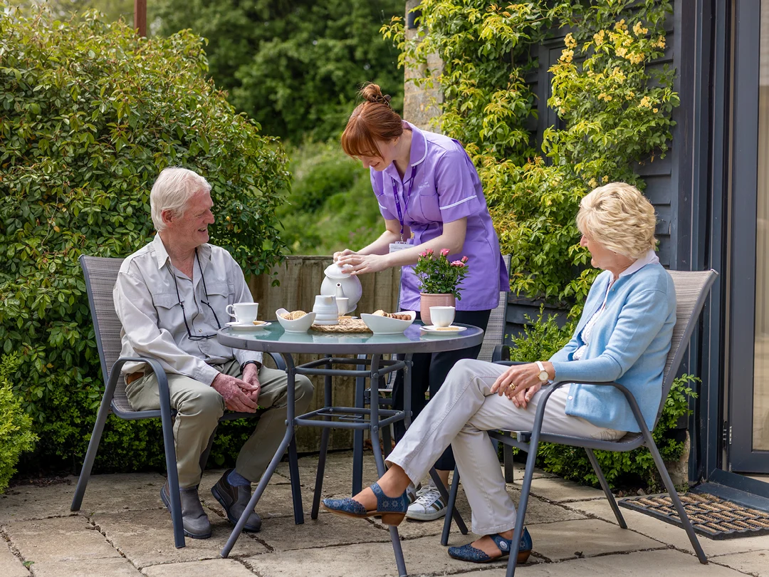 A carer pours tea for an elderly man and woman sitting at a patio table outdoors, surrounded by greenery, enjoying a peaceful afternoon together.