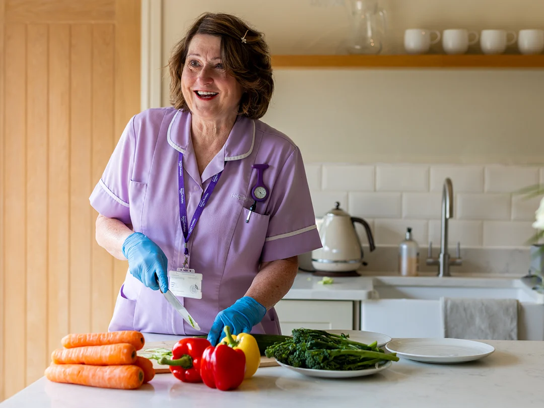 A smiling woman in a purple uniform and blue gloves slices vegetables, including peppers and carrots, on a kitchen worktop. A kettle and cups are visible in the background.